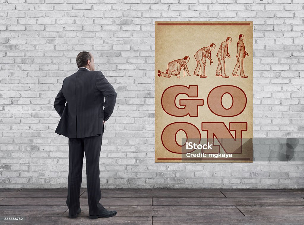 Businessman and Vintage Style “GO ON” Poster Rear view of businessman standing on wooden floor and looking at the vintage style “GO ON” poster (evolution of businessman illustration, printed on old paper: Improvement process of businessman after financial crisis. This image modeled after well-known evolution theory figure. Same businessman is changing in four steps) on the white brick wall. Adult Stock Photo