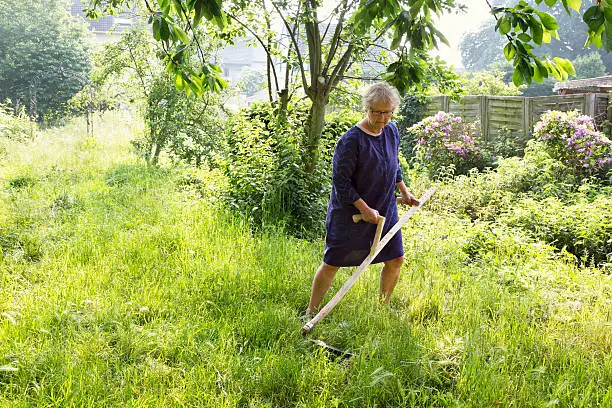 Senio woman using scythe while mowing grass in her garden