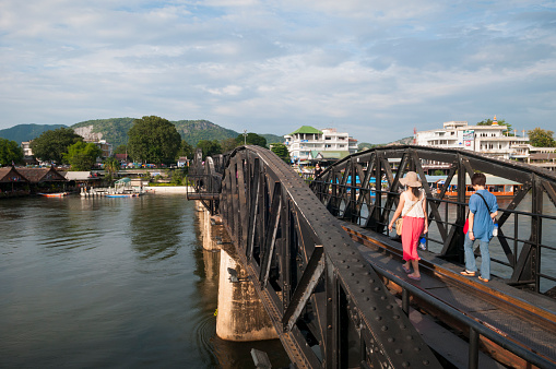 Kanchanaburi, Thailand - October 3, 2011: Two tourists walk across the famous Bridge on the River Kwai, built by Allied prisoners of war during World War II. The bridge is part of the Death Railway, so named because of the approximately 100,000 laborers, mostly conscripted Asians, who died during its construction.