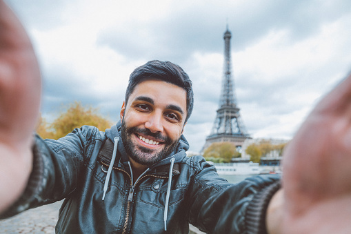 Young middle eastern man holding smart phone and taking selfie pictures. He is happy, smiling enjoying and looking at her camera at coastline of Seine river against Tour Eiffel in Paris, France. 