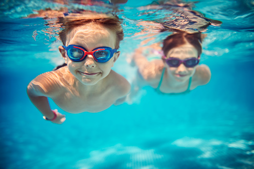 Smiling kids enjoying underwater swim in the pool towards the camera. Sunny summer day. Brother aged 5 is in the front, the sister is aged 9 and is swimming in the background. Slightly soft.