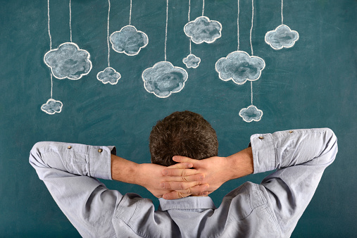 Pensive young man sitting with his face in front of a blue blackboard. He keeps hands behind head and is looking clouds. The chalkboard drawing - cloud computing concept - shows multiple clouds outlined in white and blue on a blue background. There are ten blue clouds of various sizes dangling from white strings attached to the top of the illustration. The blue background looks like a sky.