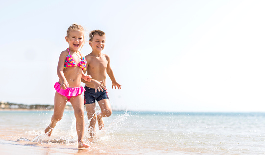 Two cheerful children having fun on the beach and running through the sea. Copy space.