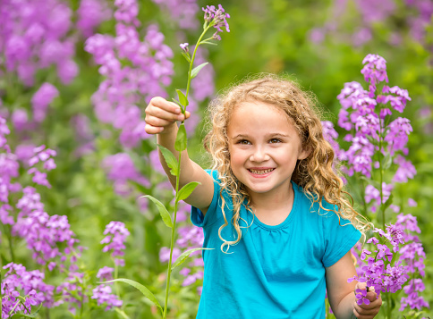 Front view of a young girl holding up and smiling at a freshly picked purple wildflower. Taken on a spring day in a rural grove of trees. The girl has curly blonde hair and is wearing a turquoise t-shirt.