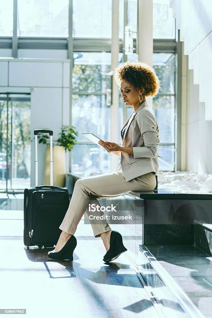Businesswoman on business travel in hotel using tablet Afro american businesswoman sitting in a hotel hall and using a digital tablet, suitcase next to her.  Business Stock Photo