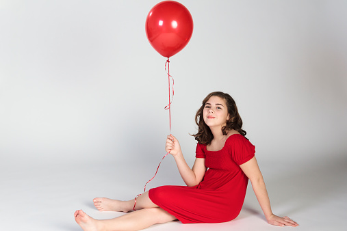 A beautiful young pre-teen girl in a red dress sitting and holding a single red balloon.