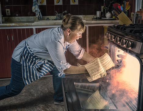 Woman  Shocked  at Oven Fire While Cooking in the Kitchen.  Smoke comes from the oven as she leans in with pot holders to reach in. Copy space.   Leica camera photograph.