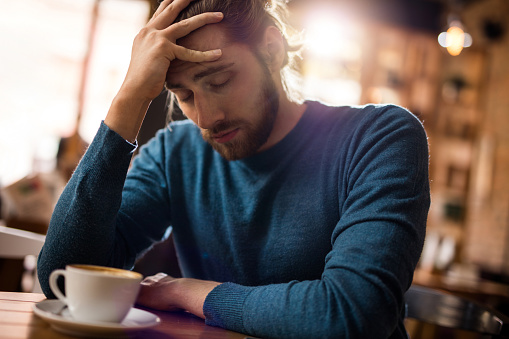 Young man with eyes closed having a headache while sitting in a cafe and holding his head in pain.