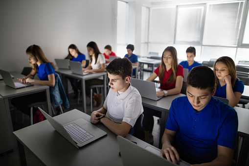 Large group of students sitting in a computer lab and using laptops. Focus is on boy in white.