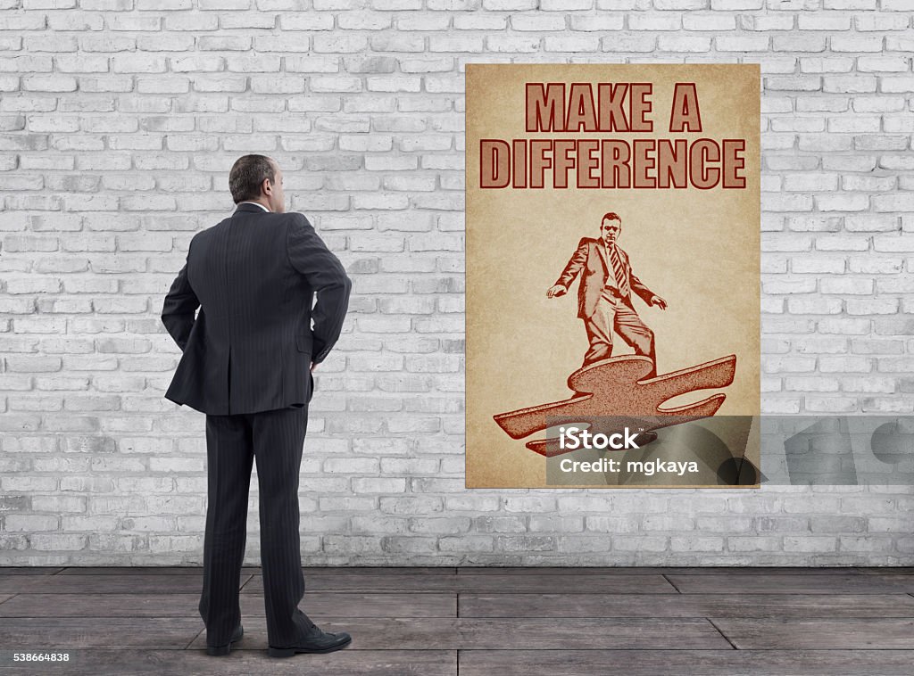 Businessman and Vintage Style “MAKE A DIFFERENCE” Poster Rear view of businessman standing on wooden floor and looking at the vintage style “MAKE A DIFFERENCE” poster (businessman flying on the piece of puzzle look like flying skateboard illustration, printed on old paper) on the white brick wall. Adult Stock Photo