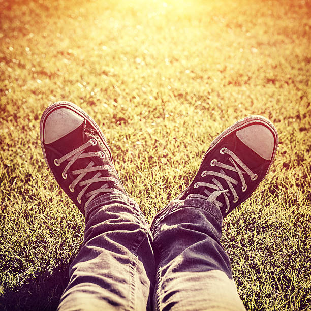 Closeup photo of lazy teen guy lying down on green grass and wearing stylish trendy gumshoes in bright yellow sunset light, body part, relaxing outdoors, fashion for hipster