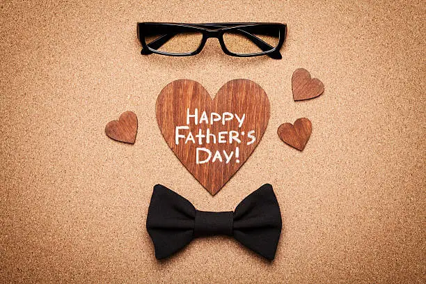 Spectacles, bowtie and wooden heart with note Happy Fathers Day, cork board background, top view, flat lay