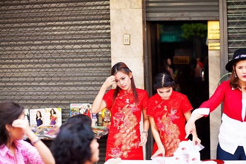 Bangkok, Thailand - February 19, 2015: Three pretty Thai girls are selling cosmetics at chinese new year. Girls are behind a table standing in Yaowarat Road of Chinatown. Two girls are wearing red chinese dress. Some people are passing scene.