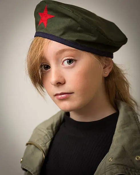 Studio portrait of a teenage ginger girl with long hair wearing a Che revolution barret hat and a green army jacket isolated against a grey background.
