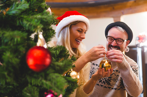 Two friends can be seen decorating the Christmas tree together, they are adding baubles to the tree and laughing together.