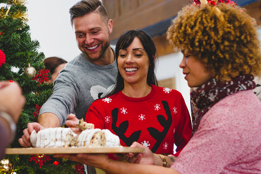 A group of friends are spending time together. A woman is holding some cake on a chopping board and all the friends are digging in and enjoying themselves. They are wearing festive, Christmas jumper.