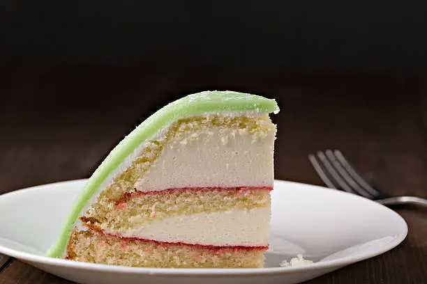 An extreme close up horizontal photograph of a slice of traditional Swedish Princess Cake. It consists of alternating layers of white cake, and pastry cream separated by thin layers of jam then it is topped with a green layer of green marzipan. The cake is isolated on black providing plenty of room for copy space.