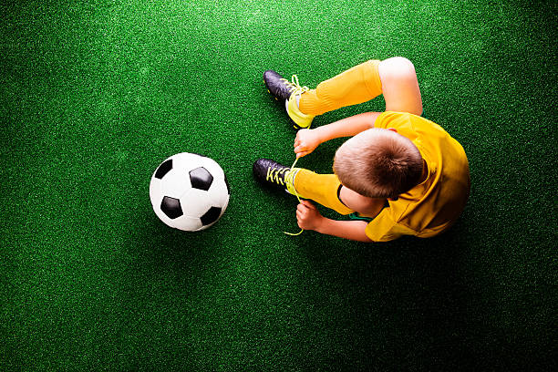 Unrecognizable little football player with soccer ball tying shoelaces, against artificial grass. Studio shot on green grass.