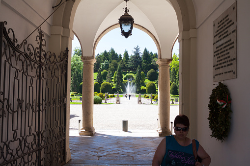 Varese, Italy - May 7, 2012: Passing gate and arcade to garden of palace Palazzo Estense in Varese at springtime. A woman is leaving garden and park. In background are people and fountain.