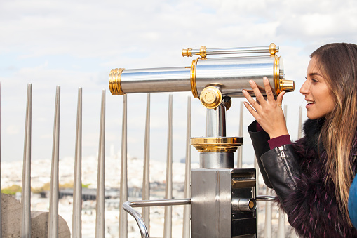 Side view of young woman standing on Arc de triomphe and using a telescope. Wears fur coat. Looking at view. Telescope in gold and silver. Cloudy sky and a railing on background.