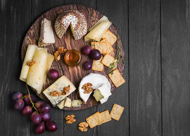 Assorted cheeses on round wooden board plate Camembert cheese, grated bark of oak, hard cheese slices, walnuts, grapes, crackers, bread, thyme, dark black wood background, top view