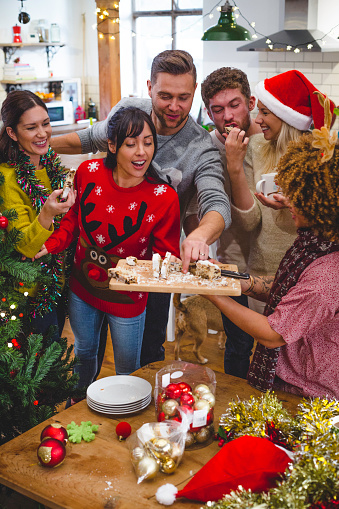A group of friends are spending time together at Christmas time. A woman is holding some cake on a chopping board and all the friends are digging in and enjoying themselves.