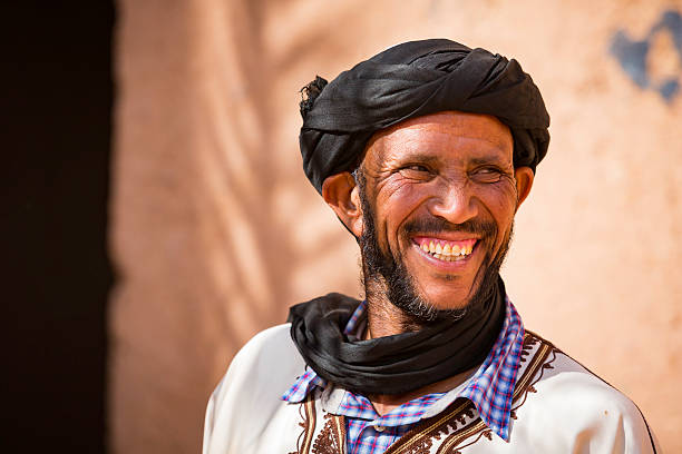 Zagora, Morocco - May 1, 2016: Moroccan mature man with beard and smiling face, wearing turban and traditional dress,  poses against the mud wall in front of an ancient, historic  Kasbah Oulad Othmane fortress