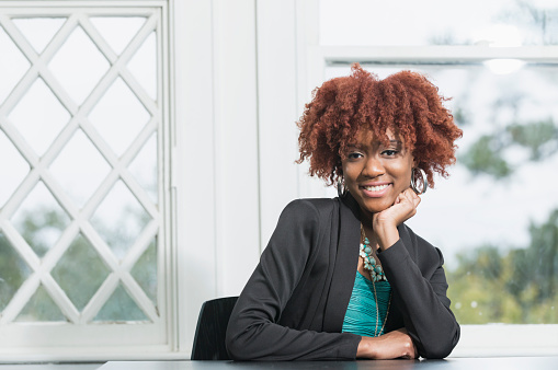 A young African American woman with an afro, stylishly dressed in a green dress and black jacket, sitting in an office boardroom, leaning her chin on her hand, smiling at the camera.