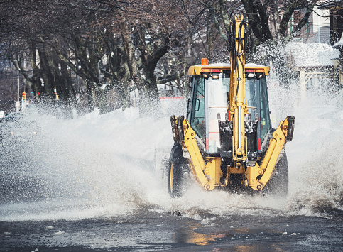 A backhoe charges through localized flooding on a city street.