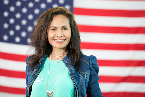 Beautiful brunette woman standing in front of the american flag wearing a denim jacket and a teal shirt.