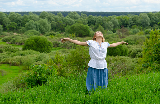 Happy girl with outspread in side hands stands on background of field with bushes and looks up at sky