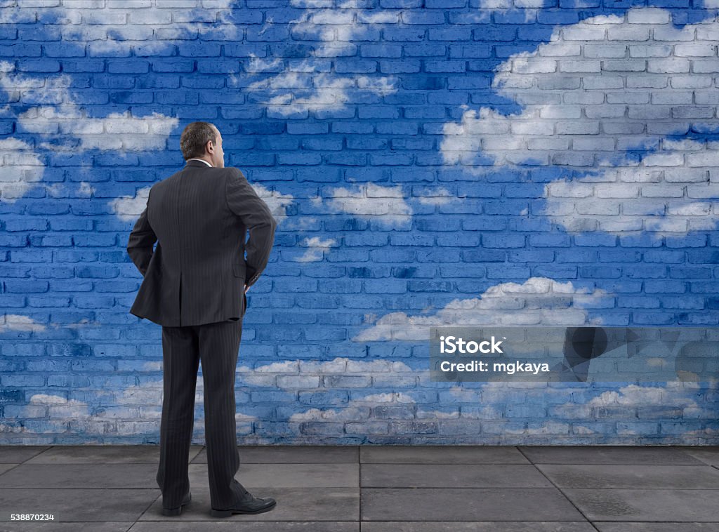 Businessman and Blue Sky with Clouds Painting on the Wall Rear view of businessman standing on sidewalk and looking at the blue sky with clouds painting on the white brick wall. Adult Stock Photo