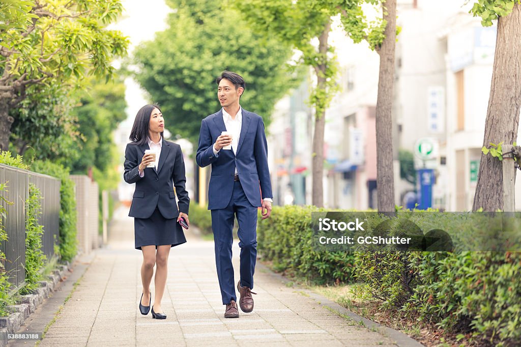 Working before even the actual office work starts Two Japanese office executives with coffee mugs walking along the street near their office and having casual conversation. Walking Stock Photo