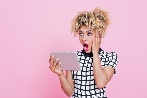 Studio portrait of shocked afro american young woman using a digital tablet. She is wearing white and black grid check playsuit. Studio portrait, pink background.