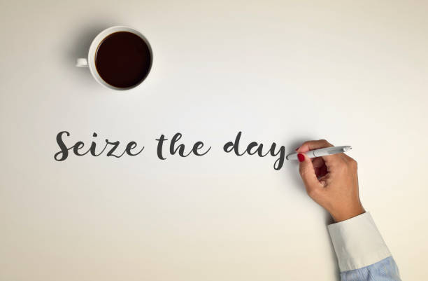 high-angle shot of a cup of coffee on an off-white surface and a young woman writing the text seize the day with a pen