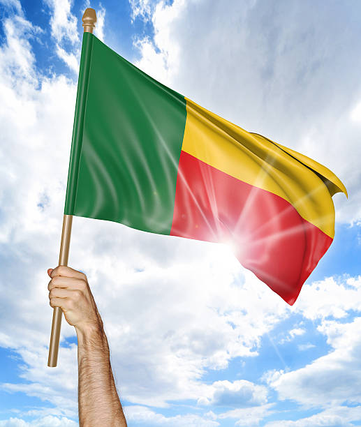 Person holding the national flag of Benin high in the air against a bright sky and sun rays.