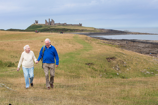 Two happy senior adults smile and hold hands as they enjoy a stroll through the countryside by the coast. The sea and a castle can be seen in the background.