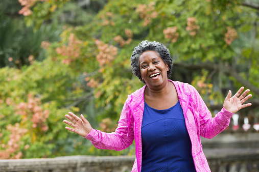 A senior, African American woman at the park, waving and talking to the camera, with a happy grin on her face.