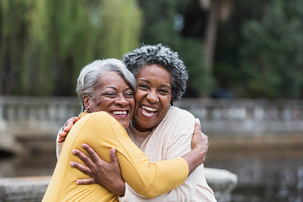 Senior black women embracing Friends hugging at the park. The two senior, African American women are standing cheek to cheek, embracing each other with big smiles on their faces, looking at the camera. cheek to cheek photos stock pictures, royalty-free photos & images