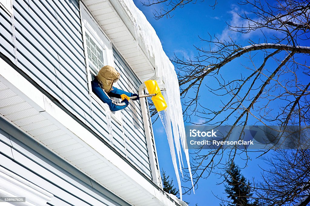 Mature Woman Leans Out Window Breaking Icicles Using a Shovel. Mature Woman Leans Out a second story Window Breaking Icicles Using a yellow Shovel.   Blue sky and copy space.  Leica camera photograph. 2015 Stock Photo