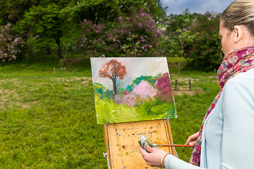Side view of a female painter working on a sketchbook painting a garden scene with flowers and outdoors cleaning off her paintbrush with a rag in a forest