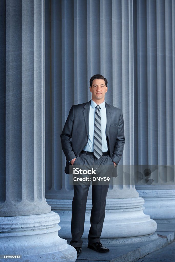 Lawyer Or Banker Standing Next To Large Columns A lawyer, banker, or businessman standing among large columns. His jacket is open and his hands are in his pockets as he looks directly at the camera. This full length portrait leaves room for copy at the top of the image.  A cool color scheme dominates the scene. Lawyer Stock Photo