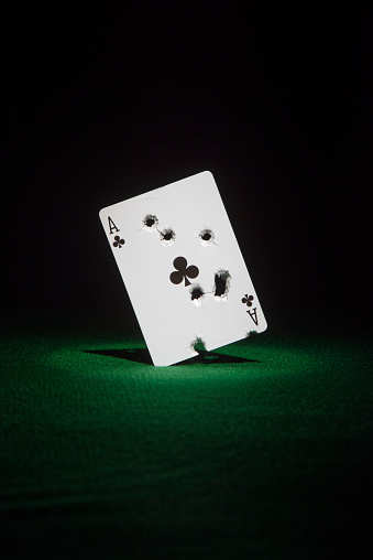 Ace of clubs with multiple bullet hole shots on poker table green, black background.