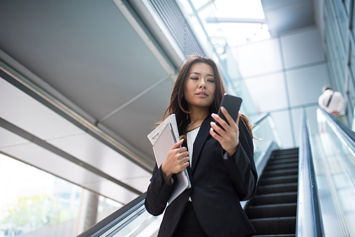 Young japanese businesswoman text messaging on escalator in front of office building. She is holding digital tablet and newspaper in one hand and she is using mobile phone with other hand.