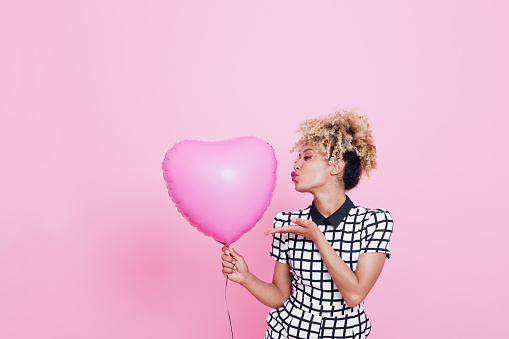 Portrait of beautiful afro american young woman wearing grid check playsuit, standing against pink background and holding Pink Heart Balloon.