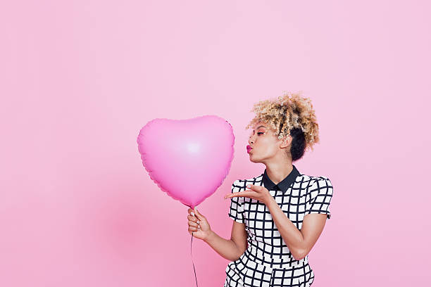 Young woman with big pink heart Portrait of beautiful afro american young woman wearing grid check playsuit, standing against pink background and holding Pink Heart Balloon. blowing a kiss stock pictures, royalty-free photos & images