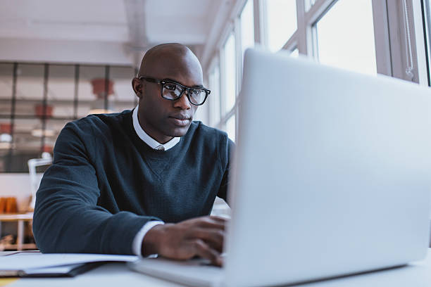 Young businessman working on his laptop Young businessman working on his laptop in office. Young african executive sitting at his desk surfing internet on laptop computer. black man laptop stock pictures, royalty-free photos & images