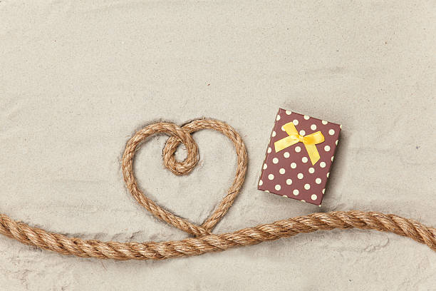 Gift box and rope in heart shape on sand background.