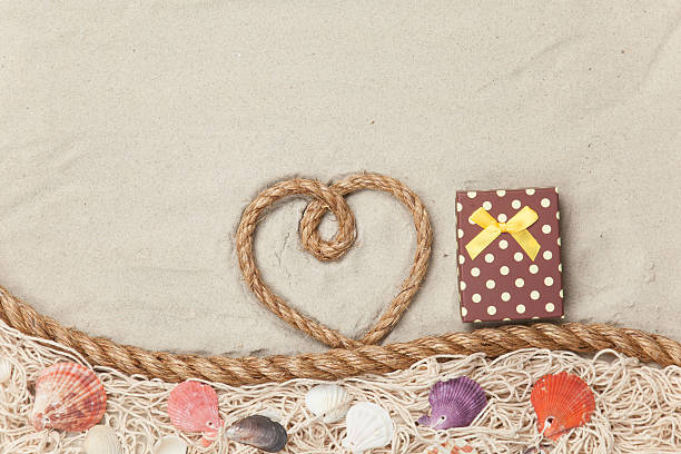 Gift box and rope in heart shape near net and shells on sand background.