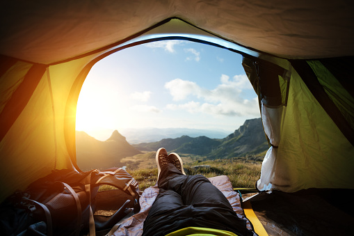 View from the tent on a beautiful mountain landscape. Traveler's legs are crossed in, backpack is laying on the left side of him. Tent is open. Beautiful mountain landscape on background, the bright sun's rays in the upper left corner of image.
